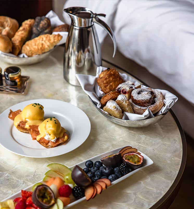 This image highlights an elegant breakfast spread at the Park Hyatt Vienna, featuring a plate of eggs benedict with smoked salmon, a selection of pastries in a silver bowl, a pitcher for coffee or tea, and a vibrant assortment of fresh fruits, including berries, figs, and passion fruit. The luxurious setup on a marble tabletop evokes a sense of indulgence and sophistication.