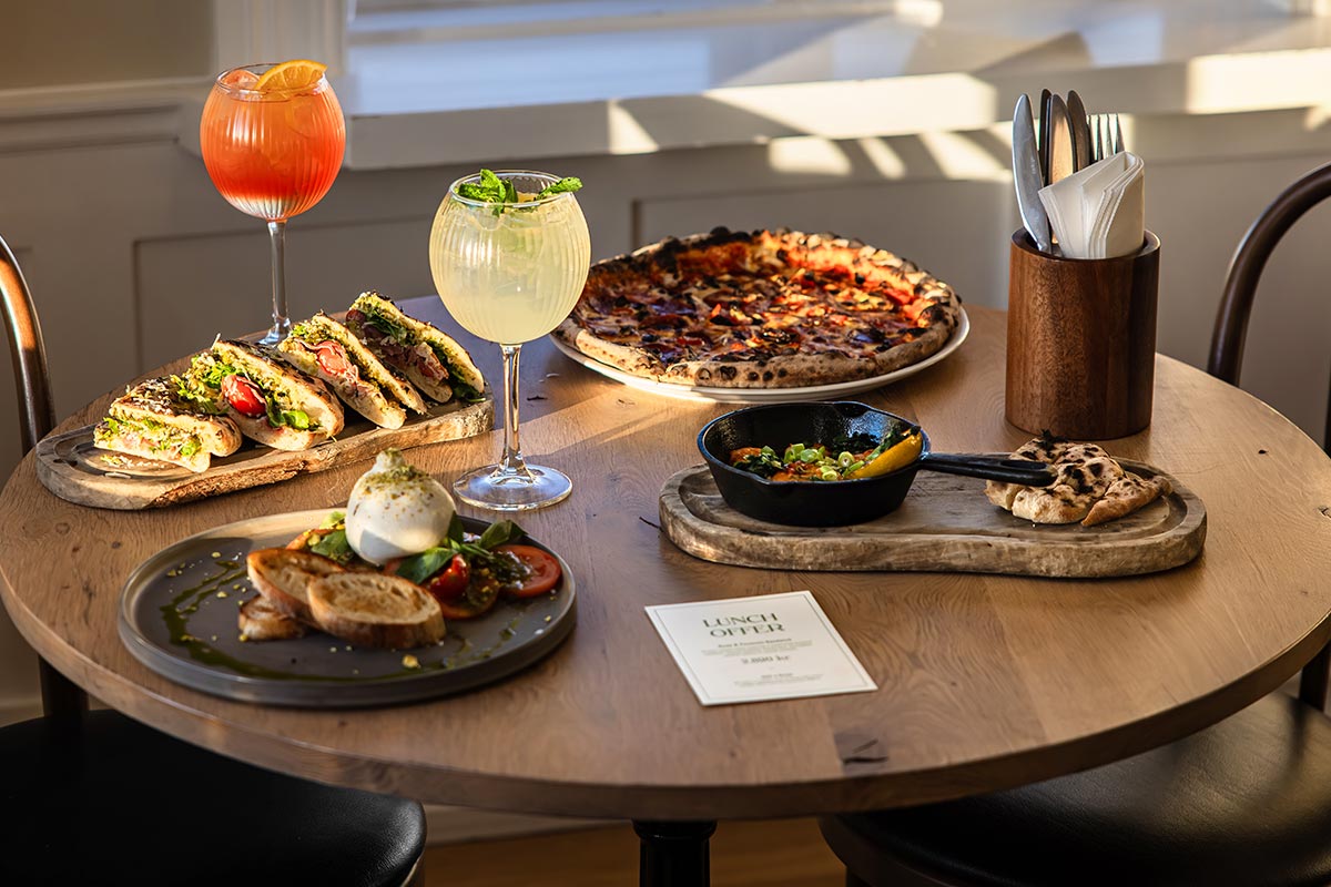 This image shows a cozy restaurant table at Raðagerði Veitingahús in Reykjavik, adorned with an array of colorful and appetizing dishes. The spread includes sandwiches, pizza, burrata with tomatoes and basil, a skillet dish, and refreshing cocktails, all illuminated by soft natural sunlight, creating a welcoming dining atmosphere.