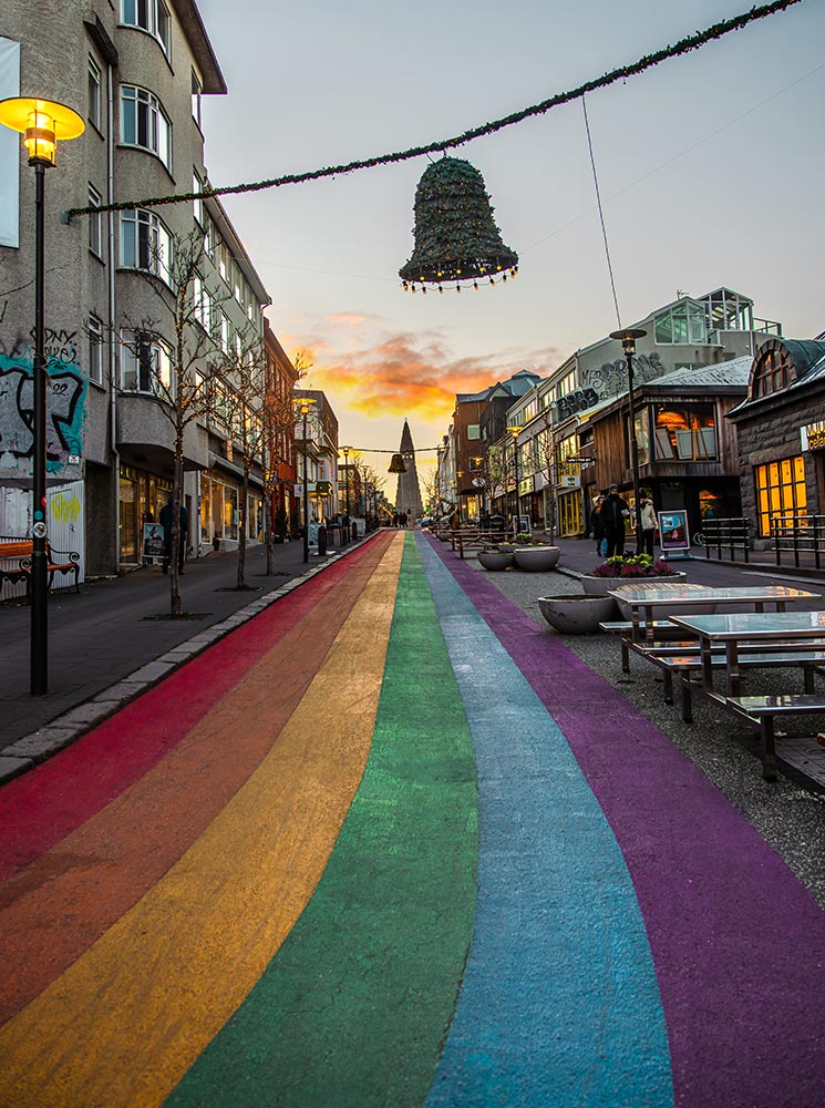 This image captures the vibrant Rainbow Street (Skólavörðustígur) in Reykjavik, leading towards Hallgrímskirkja at the end of the colorful pathway. The street is adorned with festive decorations, including a hanging bell-shaped garland, and flanked by charming shops and buildings, all illuminated by a warm sunset.