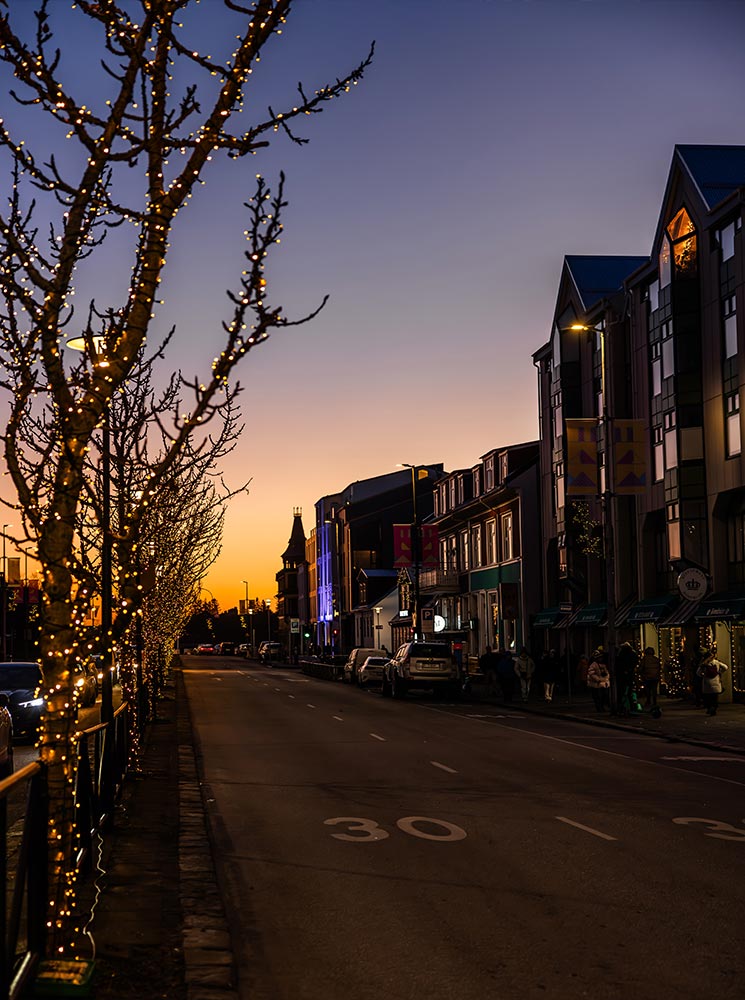 This image captures a quiet street in Reykjavik during sunset, with a vibrant orange and purple sky providing a beautiful backdrop. Trees lining the street are wrapped in warm fairy lights, adding a festive touch to the evening scene. The softly illuminated buildings and scattered holiday decorations create a cozy and charming atmosphere.