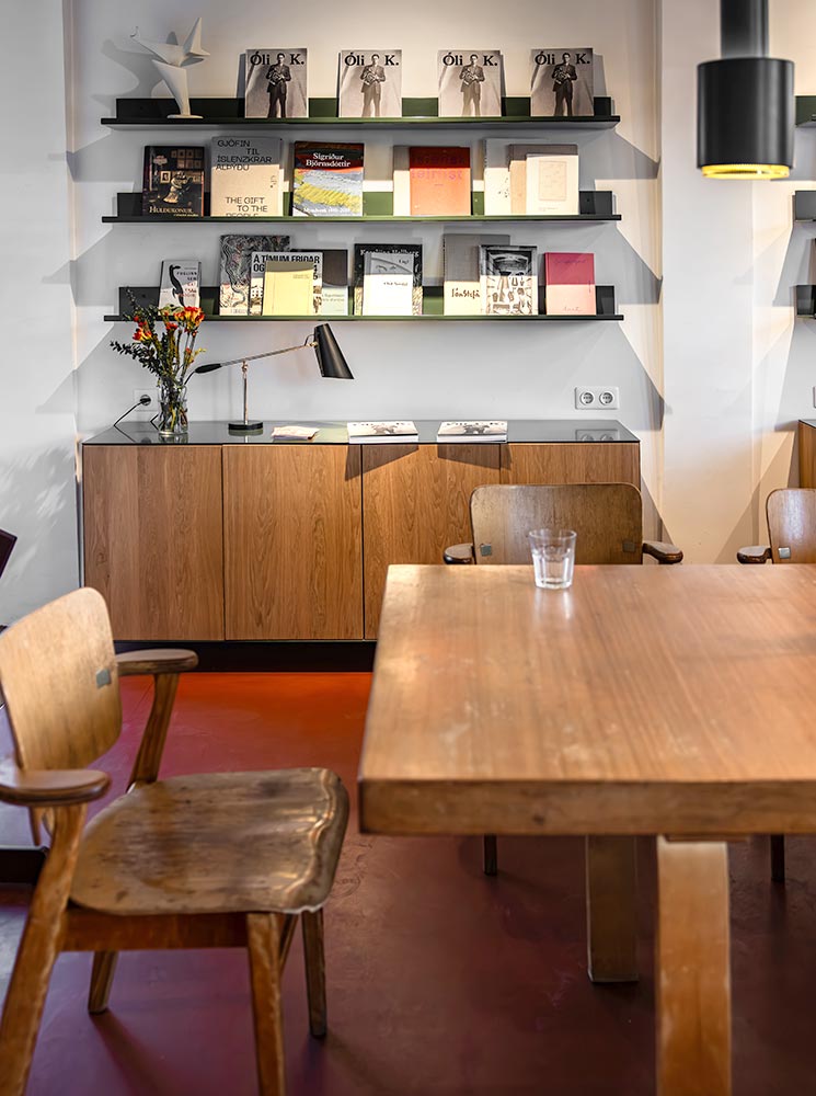 This image showcases a stylish café interior with wooden furniture and a modern aesthetic. A wall-mounted bookshelf displays magazines and books, including copies of "Óli K.," above a wooden cabinet adorned with a lamp and a vase of flowers. The cozy atmosphere is enhanced by warm lighting and a minimalist design.