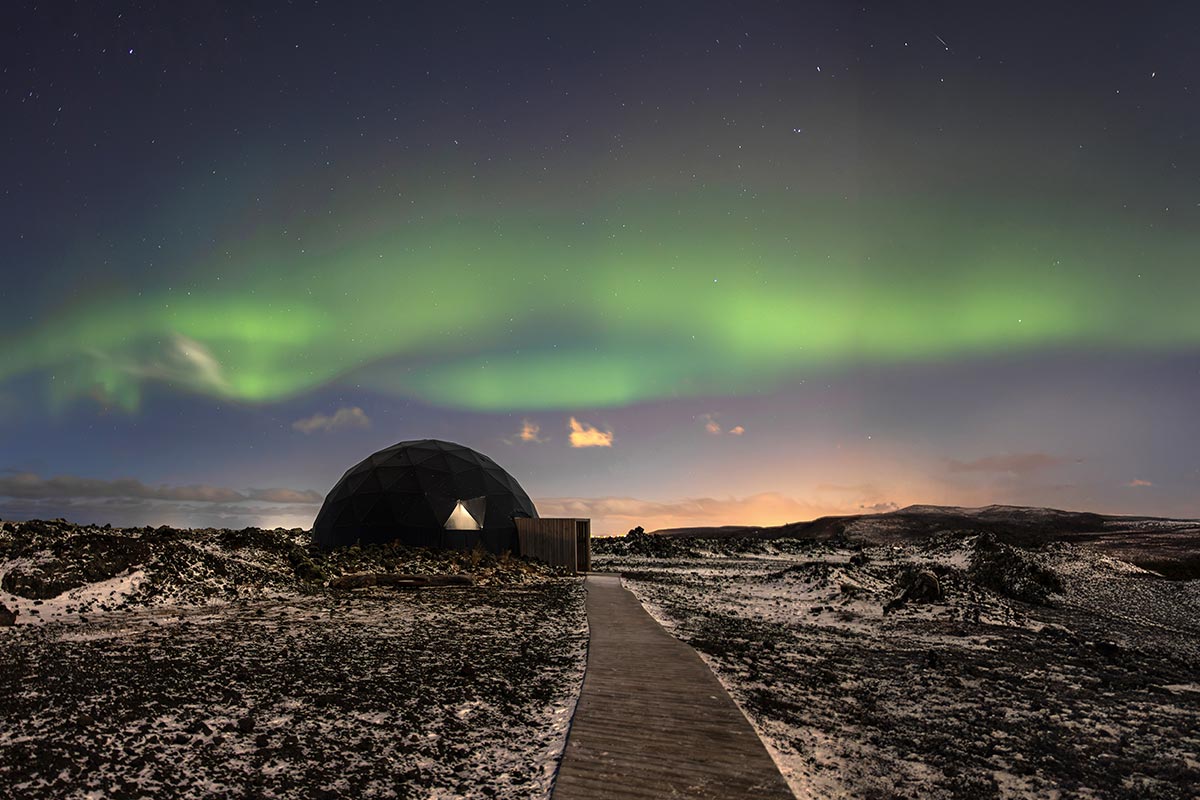 This image shows a geodesic dome structure under a vivid display of the Northern Lights (Aurora Borealis) against a starry night sky. The foreground features a wooden pathway leading to the dome, surrounded by a rugged, snow-dusted landscape.