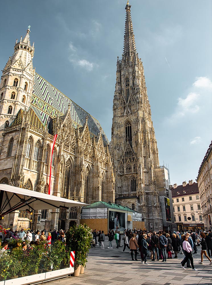 This image showcases St. Stephen's Cathedral in Vienna, with its towering Gothic spire and distinctive patterned tiled roof. The busy plaza below is filled with people, some gathered near a café with outdoor seating. The cathedral’s intricate stonework contrasts with the bustling modern scene, capturing a blend of historical and contemporary Vienna.