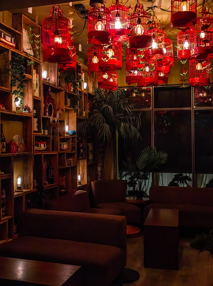 This image showcases the cozy and stylish interior of Sushi Social in Reykjavik, with a unique ambiance created by vibrant red birdcage-inspired pendant lights hanging from the ceiling. The dimly lit space features comfortable seating and lush greenery, blending a modern aesthetic with a warm, intimate atmosphere. The setting exudes sophistication, making it an inviting spot for enjoying sushi and cocktails.