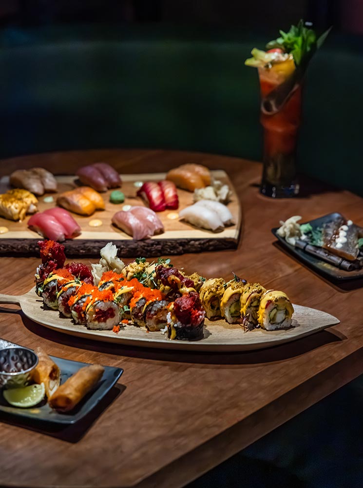 This image features an assortment of beautifully presented sushi rolls and nigiri served on wooden trays at Sushi Social in Reykjavik. The vibrant rolls are adorned with colorful toppings like fish roe and garnishes, while a cocktail sits elegantly in the background. The scene captures the essence of fine dining with a creative and fresh take on sushi.