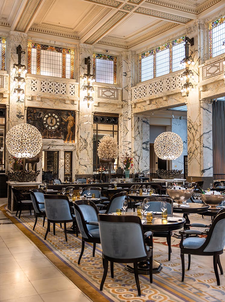 This image captures the luxurious interior of The Bank Brasserie & Bar at the Park Hyatt Vienna, showcasing its elegant decor with marble walls, stained glass windows, and large spherical chandeliers. The room is filled with neatly set tables and plush blue chairs, creating a refined dining atmosphere. The intricate ceiling design and Art Deco accents emphasize the venue's sophisticated and historic charm.