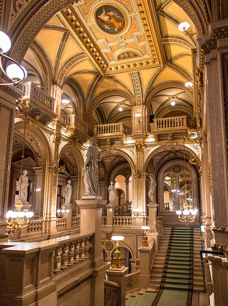 This image showcases the grand staircase inside the Vienna State Opera, surrounded by marble statues, arched ceilings, and intricate gold details. The ceiling features a beautiful fresco, and elegant chandeliers cast a warm glow over the ornate architectural elements. The luxurious setting reflects the historic grandeur and artistic richness of one of Vienna’s most iconic cultural landmarks.