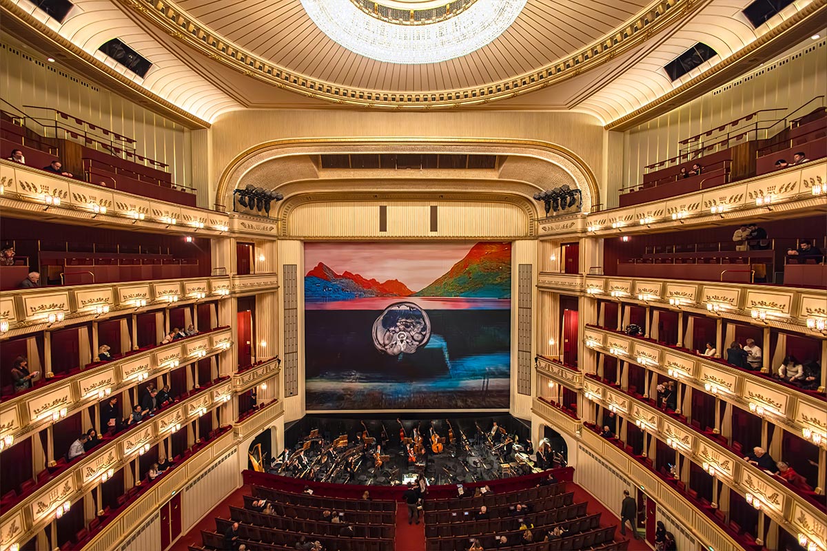 This image captures the grand interior of the Vienna State Opera, showcasing its elegant balconies with red velvet seating and gold accents under a large, radiant chandelier. The stage features a colorful, scenic backdrop of mountains and water, set for an upcoming performance. The orchestra pit is filled with musicians preparing, creating a refined and anticipatory atmosphere in this iconic venue.






