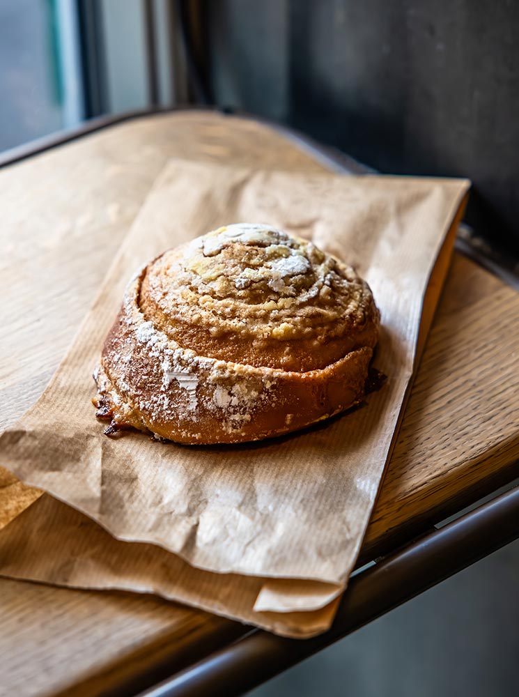 This image showcases a freshly baked cinnamon roll from Brauð & Co. in Reykjavik, resting on a simple brown paper wrap. The pastry is golden-brown, generously dusted with powdered sugar, and has a rich, flaky texture, evoking a sense of warmth and indulgence.