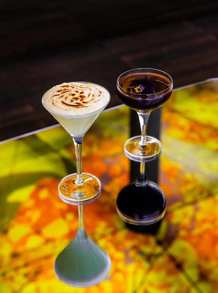 This image showcases two elegant cocktails served in martini and coupe glasses on a colorful, abstract tabletop at Das LOFT bar in Vienna. One drink appears creamy with a caramelized frothy top, while the other is dark with a delicate flower garnish. The vibrant reflections on the table enhance the artistic presentation, adding to the chic, upscale atmosphere.