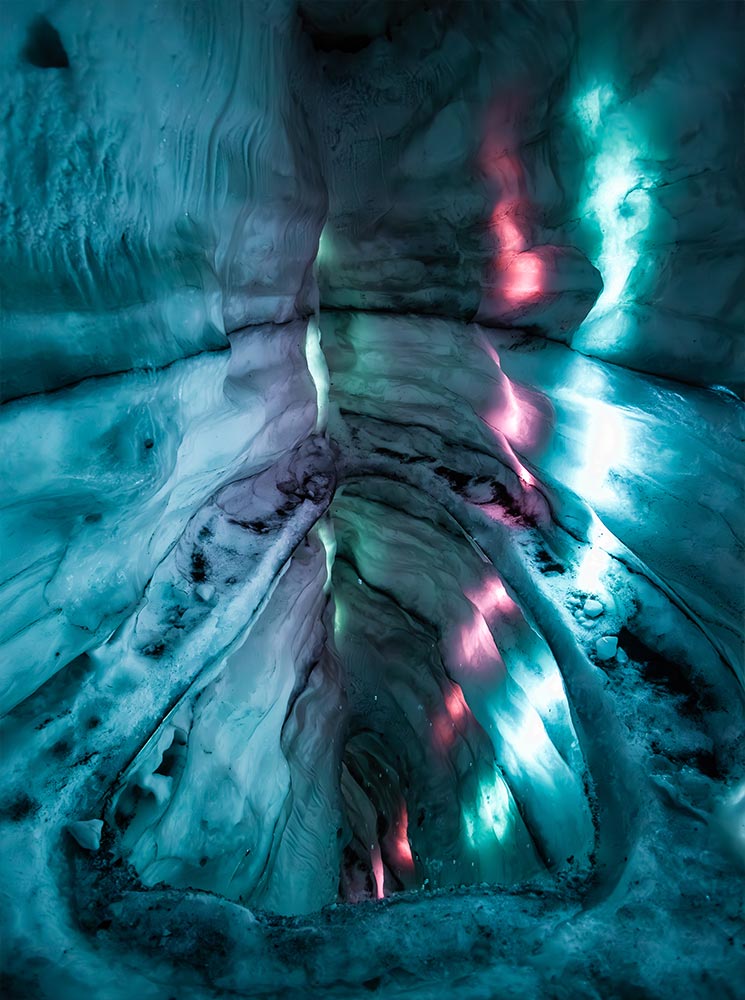 This image depicts the mesmerizing ice cave exhibit at the Perlan Museum in Reykjavik, featuring smooth, sculpted walls of ice illuminated by vibrant blue, green, and pink lights. The textured surfaces and glowing colors create an ethereal, otherworldly atmosphere, showcasing the beauty of Iceland's icy landscapes in an indoor setting.