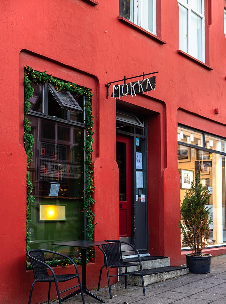 This image shows the exterior of Mokka Kaffi, a cozy café in Reykjavik, featuring a vibrant red facade decorated with festive greenery around the windows. A small outdoor seating area with black chairs and a table adds a welcoming touch, complementing the café’s rustic charm.