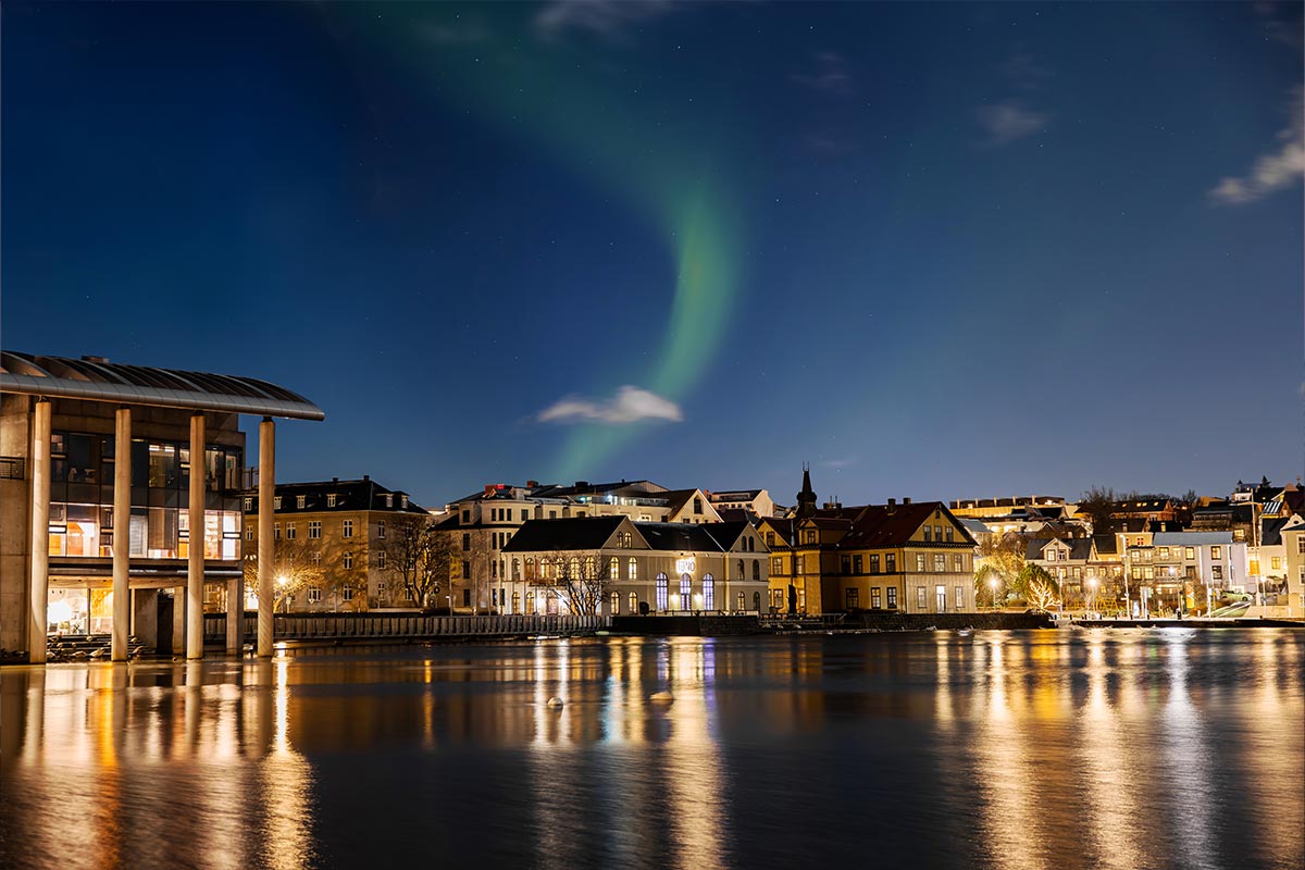 This image depicts a stunning night view of Reykjavik, with the Northern Lights creating a green ribbon of light in the starry sky above the city. The reflection of the lights and surrounding buildings shimmers on the calm waters of the lake, highlighting the charm of the city's architecture. The combination of natural beauty and urban warmth makes for a captivating scene.