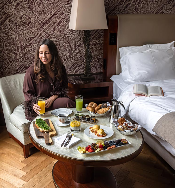 This image depicts a cozy in-room breakfast scene at the Park Hyatt Vienna. A woman sits beside a table laden with a luxurious spread, including fresh pastries, eggs benedict, fruit, toast with avocado, and juices. The room’s elegant decor, with rich textures and soft lighting, adds to the relaxed and indulgent atmosphere.