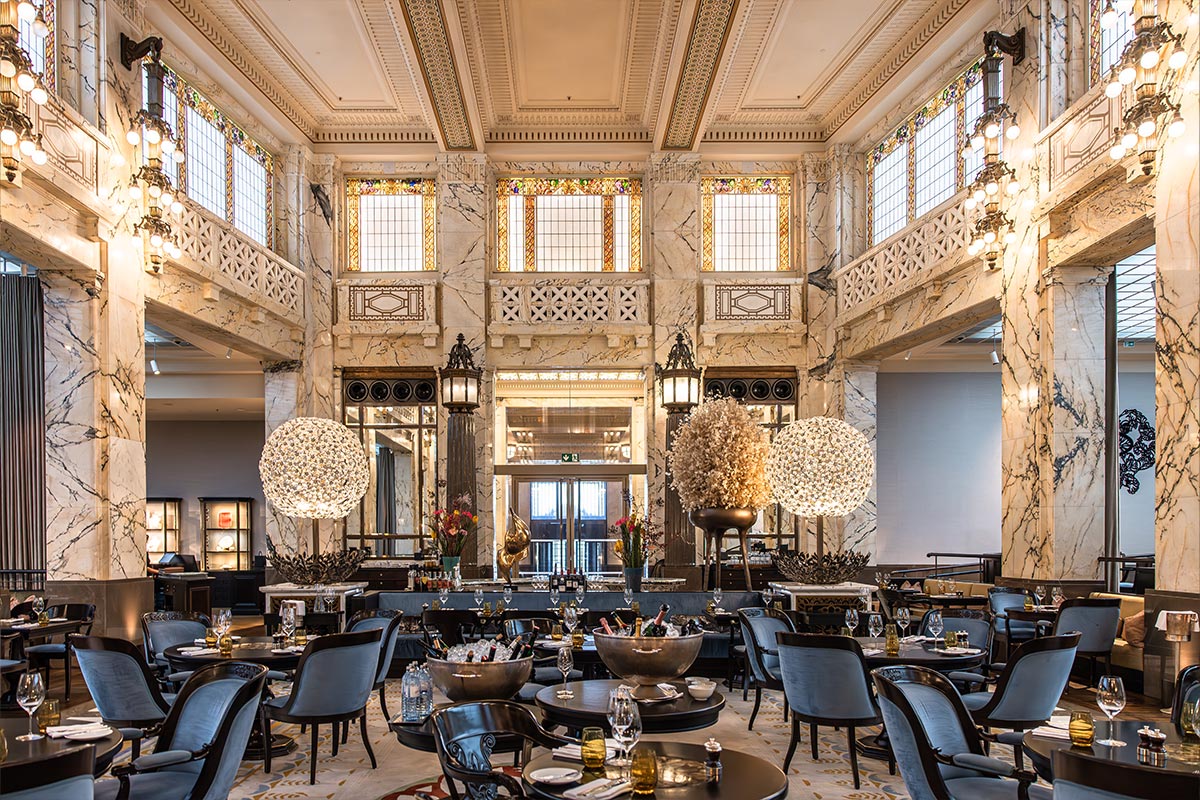 This image showcases an elegant interior of the Park Hyatt Vienna hotel, featuring a grand dining area with marble pillars, intricate stained glass windows, and ornate chandeliers. The space is adorned with spherical light fixtures, decorative floral arrangements, and plush seating, creating a sophisticated and luxurious ambiance.