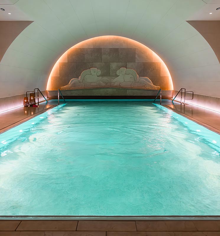 This image shows the luxurious indoor pool at the Park Hyatt Vienna, set within a serene arched room. The pool is softly illuminated with a calming glow from the surrounding lights, highlighting an elegant relief artwork on the back wall, featuring reclining figures, which adds to the tranquil ambiance.