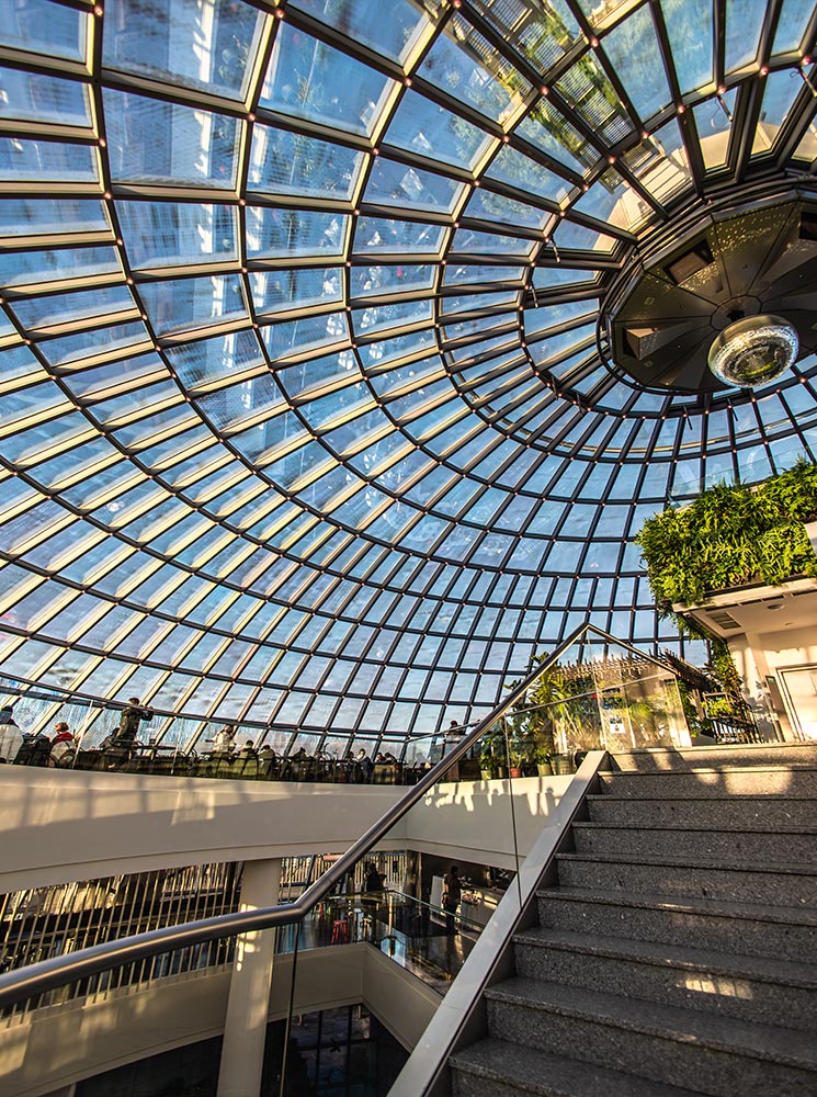 This image captures the striking glass dome of the Perlan Museum in Reykjavik, showcasing its intricate, sunlit lattice structure. The interior features modern staircases and railings, surrounded by lush greenery and natural light streaming through the ceiling, creating a vibrant and airy atmosphere. Visitors are visible enjoying the panoramic views from the upper levels.