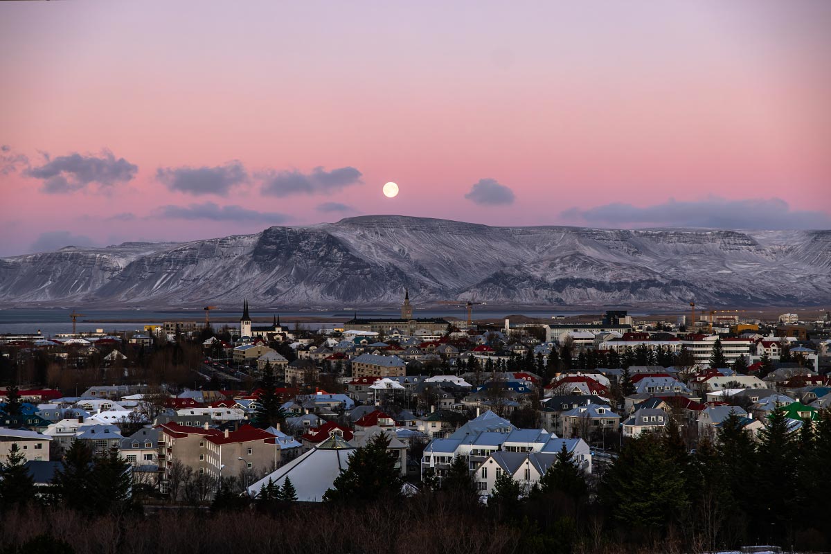 This image captures a serene view of Reykjavik from the observation deck at the Perlan Museum. The cityscape, featuring colorful rooftops and iconic church spires, is framed by a snow-capped mountain range in the distance. A full moon rises against a pastel pink and purple twilight sky, creating a peaceful and picturesque scene.