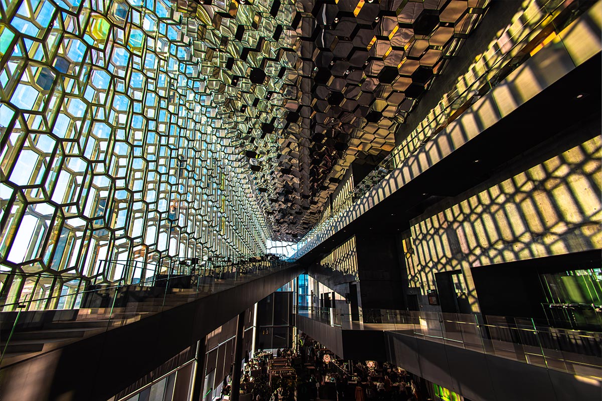 This image captures the stunning interior of Harpa Concert Hall in Reykjavik, with its signature honeycomb-shaped glass panels that reflect light in a beautiful, kaleidoscopic pattern. The interplay of sunlight through the glass creates striking shadows across the floors and walls, highlighting the building's modern and architectural grandeur. The open space and glass facades offer a dramatic, airy feel.