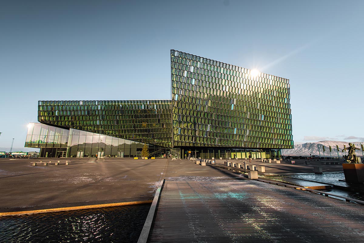 This image features Harpa Concert Hall in Reykjavik, an architectural masterpiece with its distinctive glass facade reflecting hues of green and blue. The geometric patterns of the building catch the sunlight, creating a dazzling display against a clear sky. The surrounding plaza and tranquil waters further enhance the modern and scenic ambiance of this iconic landmark.