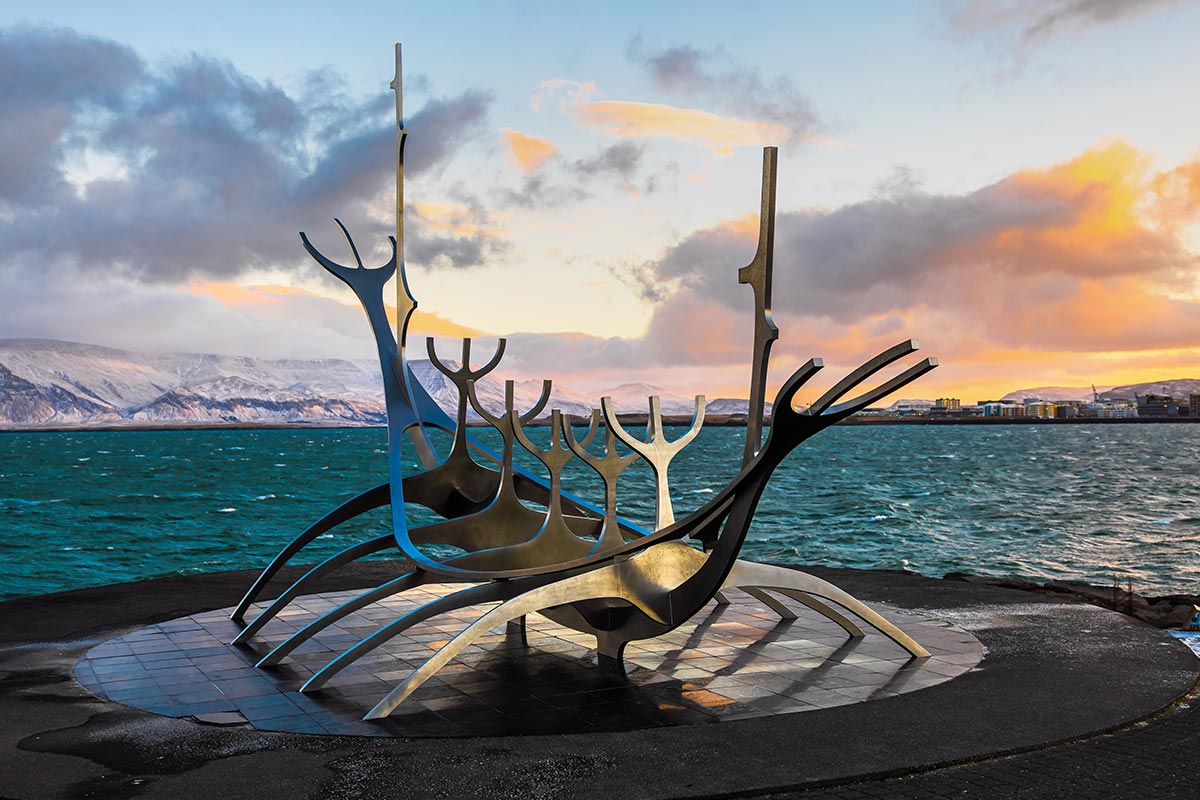 This image captures the Sun Voyager sculpture in Reykjavik, Iceland, with its sleek, metallic design resembling a viking ship set against a stunning backdrop of the ocean and snow-capped mountains. The warm hues of the sunrise or sunset add a dramatic contrast to the icy blue waters, creating a moment of serene beauty and symbolism.