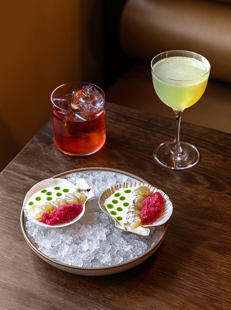 This image features a sophisticated dining setup with two elegant cocktails—a vibrant red Negroni garnished with herbs and a light green drink in a coupe glass. Accompanying the drinks is a beautifully plated dish of fresh seafood served in scallop shells on a bed of crushed ice, garnished with bright pink roe and green dots of sauce. The presentation highlights a refined culinary experience.