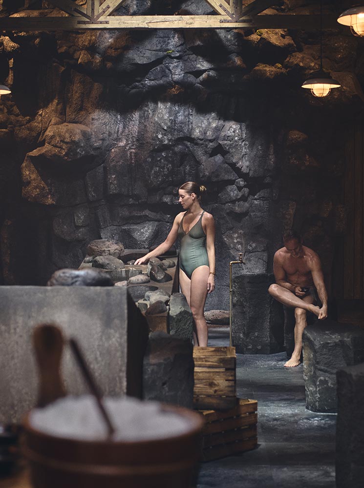 This image showcases the serene interior of the Sky Lagoon's 7-Step Ritual in Reykjavik, highlighting a rustic, spa-like setting with natural stone surroundings and soft lighting. A woman in a swimsuit stands near a stone basin, immersed in the tranquil ambiance, while another person relaxes nearby. The atmosphere reflects a blend of Icelandic tradition and modern wellness practices, creating a deeply rejuvenating experience.