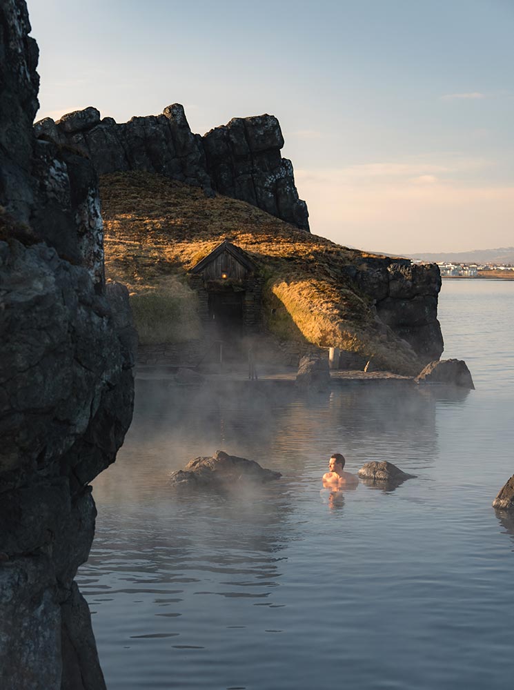 This image highlights the tranquil beauty of the Sky Lagoon in Reykjavik, featuring a lone bather enjoying the warm geothermal waters amidst natural rock formations. A small, traditional Icelandic turf house is nestled into the hillside, adding a rustic charm to the serene landscape. The soft light and rising steam create a peaceful, rejuvenating atmosphere, showcasing Iceland's unique spa culture.