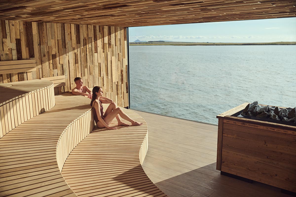 This image showcases the serene sauna at Sky Lagoon in Reykjavik, with its sleek wooden interior and a panoramic window offering a stunning view of the ocean. Guests are seen relaxing on the curved wooden benches, enjoying the calming natural light and tranquil surroundings. The minimalist design and breathtaking scenery create a perfect blend of comfort and Icelandic nature.