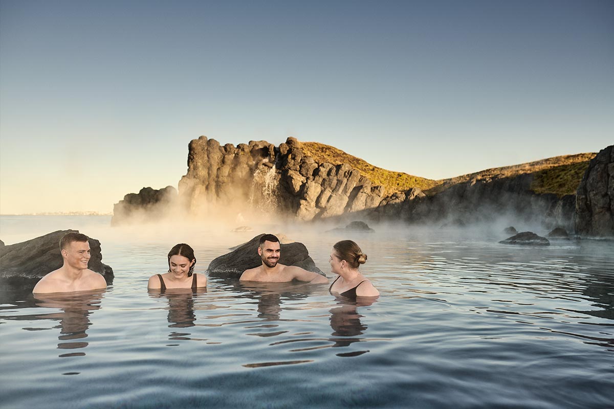 This image showcases a serene moment at the Sky Lagoon in Reykjavik, where a group of people relax in the warm geothermal waters surrounded by rugged coastal cliffs. Steam gently rises from the water under a clear blue sky, enhancing the tranquil and rejuvenating atmosphere. The natural setting, combined with the lagoon's peaceful ambiance, highlights Iceland's unique geothermal experiences.