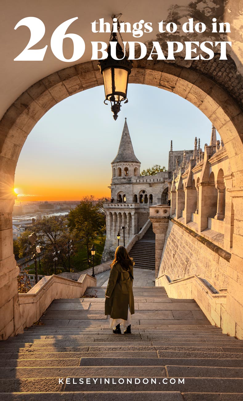 This image is a travel guide cover titled "26 Things to Do in Budapest" by Kelseyinlondon.com. It features a woman standing on steps, facing the Fisherman's Bastion in Budapest, with an arch framing the view. The sun sets in the background, casting a warm glow over the historic architecture and the surrounding landscape.