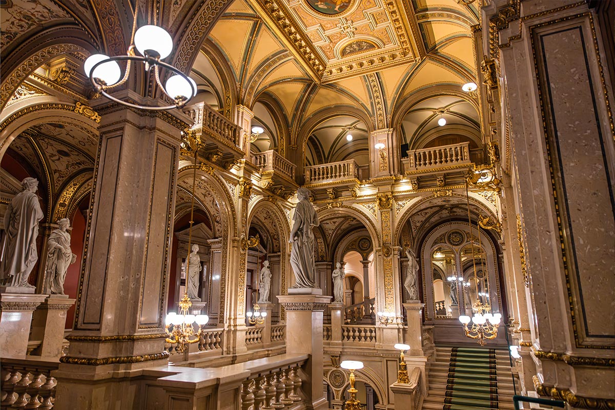 This image captures the opulent interior of the Vienna State Opera, showcasing its grand staircase, ornate arches, and classical statues bathed in warm lighting. The intricate ceiling design features rich gold accents and detailed frescoes, adding to the luxurious atmosphere of this historic venue.






