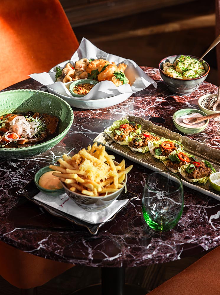 This image displays a variety of dishes at Chez Bernard in Vienna, arranged on a dark marble table. The spread includes golden fries with dipping sauce, a plate of tacos topped with chili and herbs, a green bowl of ramen-like noodles, a dish of dumplings with sauce, and a creamy potato side garnished with chives. The vibrant colors and textures of the food create a visually appealing and appetizing display.