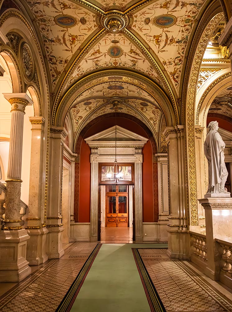 This image features an opulent corridor inside the Vienna State Opera, showcasing an arched ceiling adorned with intricate frescoes and gilded details. The pathway is lined with marble columns and leads to a warmly lit doorway framed by rich red walls, with a classical statue standing to the side. The elegant decor reflects the historic and cultural significance of this iconic venue.