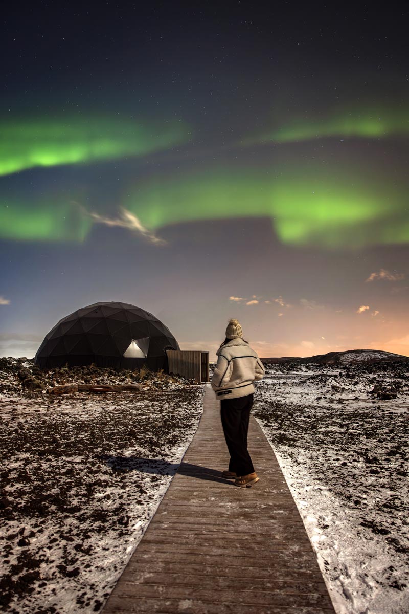 This image captures a person walking on a wooden path toward a geodesic dome under a vibrant display of the Northern Lights at the Aurora Basecamp near Reykjavik. The snow-dusted ground and the glowing green aurora overhead create a magical, otherworldly atmosphere, blending natural beauty with the serene structure in the foreground.