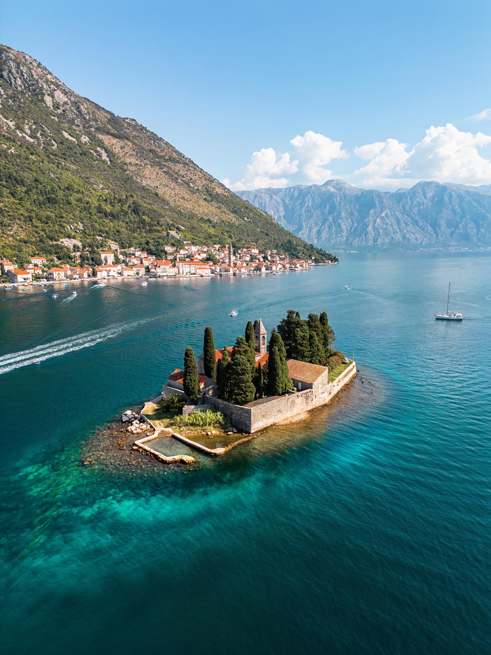 An aerial view of Saint George Monastery, a small island in the Bay of Kotor, Montenegro, surrounded by turquoise waters, with lush cypress trees and a stone church, set against the backdrop of Perast and majestic mountains.