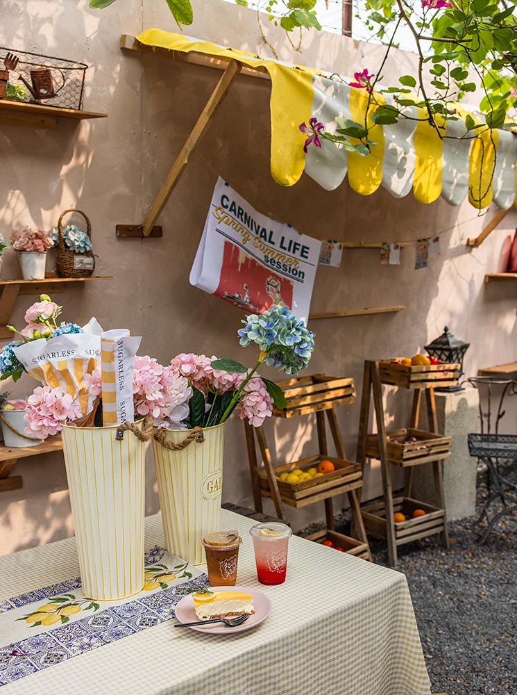 This image features a charming outdoor cafe setup with a table decorated with a lemon-patterned runner, pastel flowers in vintage-style tins, and two drinks—one coffee and one a pink soda. A slice of cheesecake sits on a pink plate, completing the inviting spread. The background includes wooden shelves and crates filled with fruit, and a playful "Carnival Life" poster hangs on the stucco wall under a yellow-striped canopy.