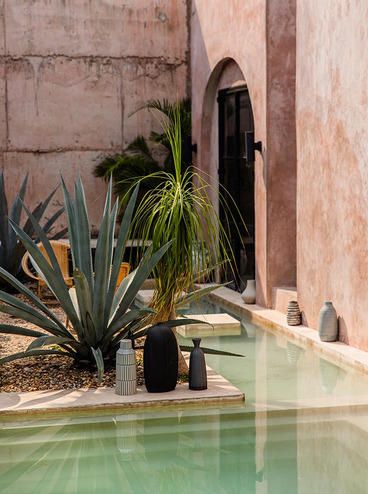 This image depicts a stylish outdoor space featuring a minimalistic desert-inspired design. A large agave plant and decorative vases are placed near a shallow reflective pool, set against earthy pink stucco walls with an arched black-framed window. The tranquil and modern aesthetic creates a serene and sophisticated atmosphere.