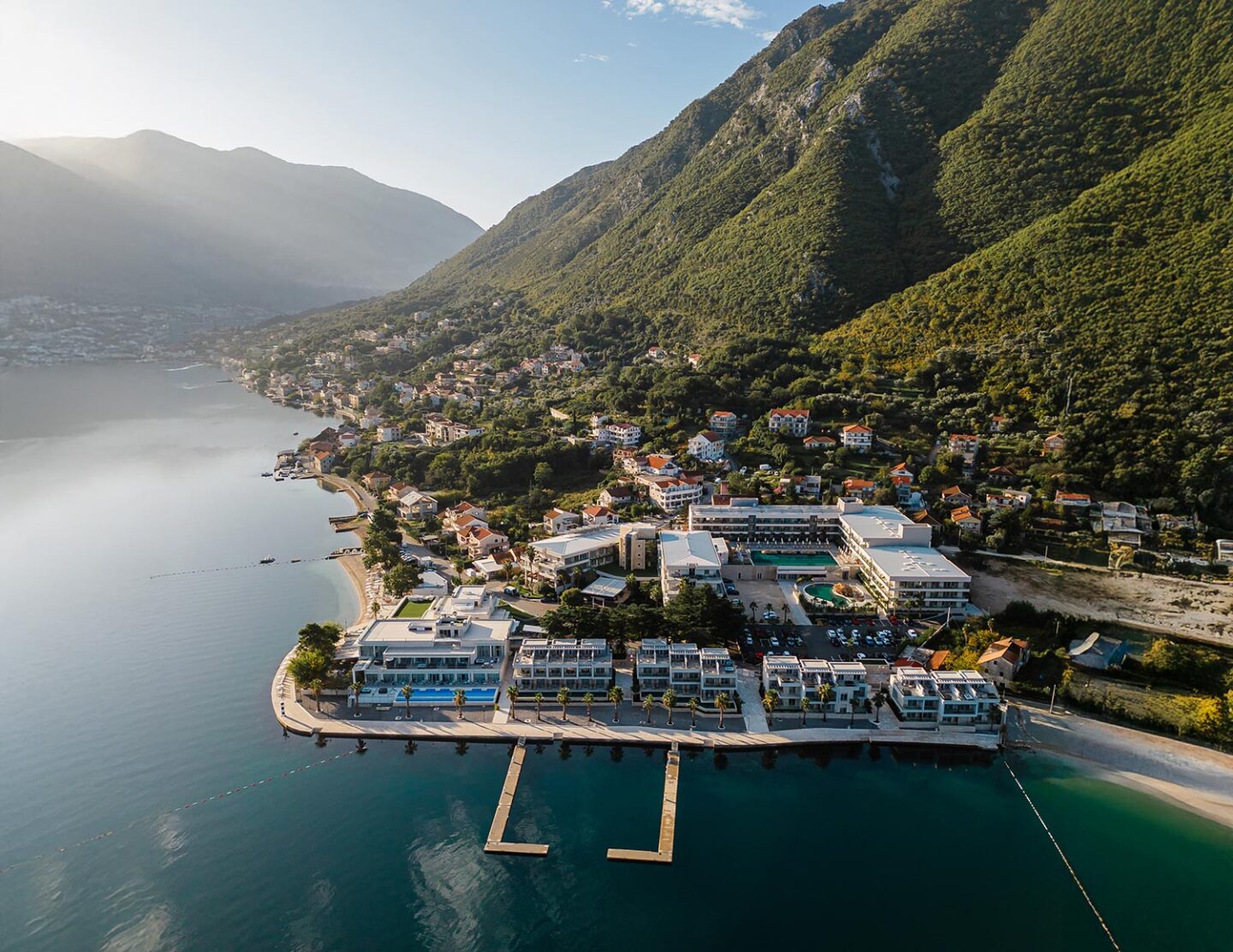 An aerial view of the Hyatt Regency Kotor Bay Resort, situated along a serene coastline with modern buildings, a dock extending into the calm bay, and lush green mountains in the background under soft sunlight.