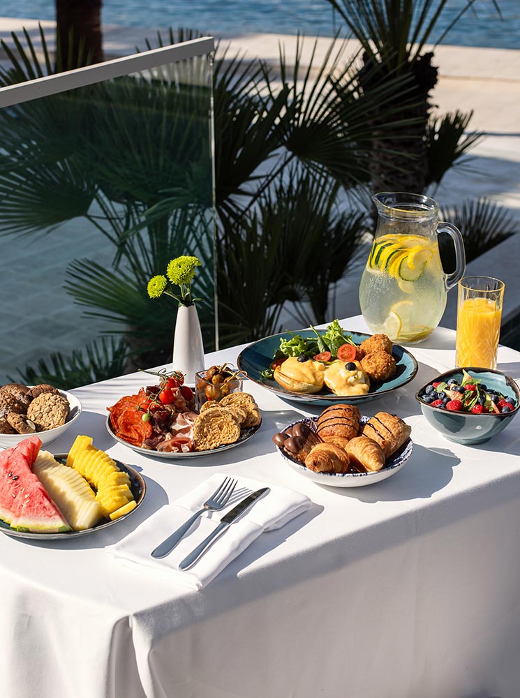 A beautifully arranged breakfast table overlooking the water, featuring a variety of dishes including fresh fruit, pastries, bread, cheese, cold cuts, salad, a bowl of mixed berries, and a pitcher of lemon water, alongside a glass of orange juice.