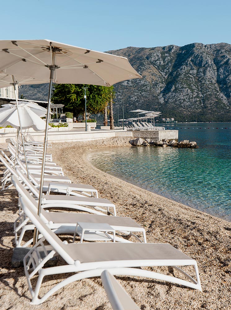 A serene private beach at the Hyatt Regency Kotor Bay Resort, featuring rows of white sun loungers and umbrellas on a pebbled shore, overlooking clear turquoise water with a backdrop of majestic mountains.