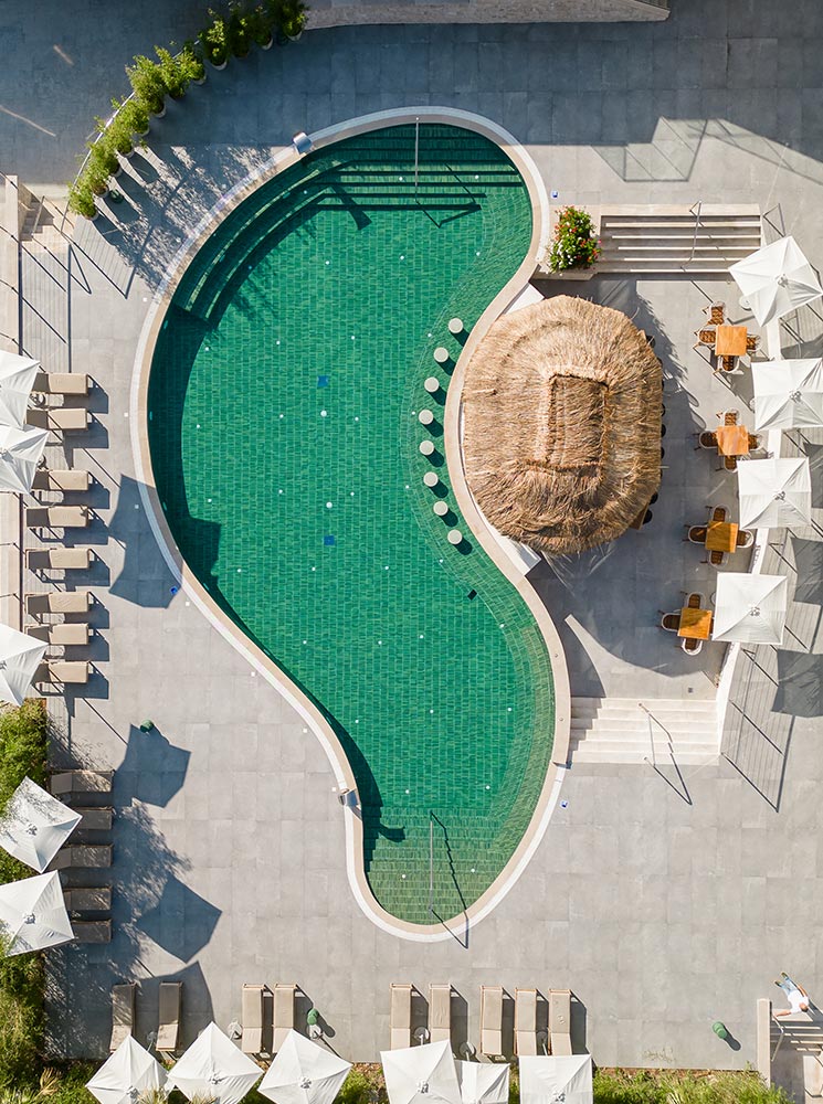 An aerial view of a uniquely shaped swimming pool at the Hyatt Regency Kotor Bay Resort, surrounded by sun loungers, umbrellas, and a thatched-roof bar, creating a relaxing poolside atmosphere.