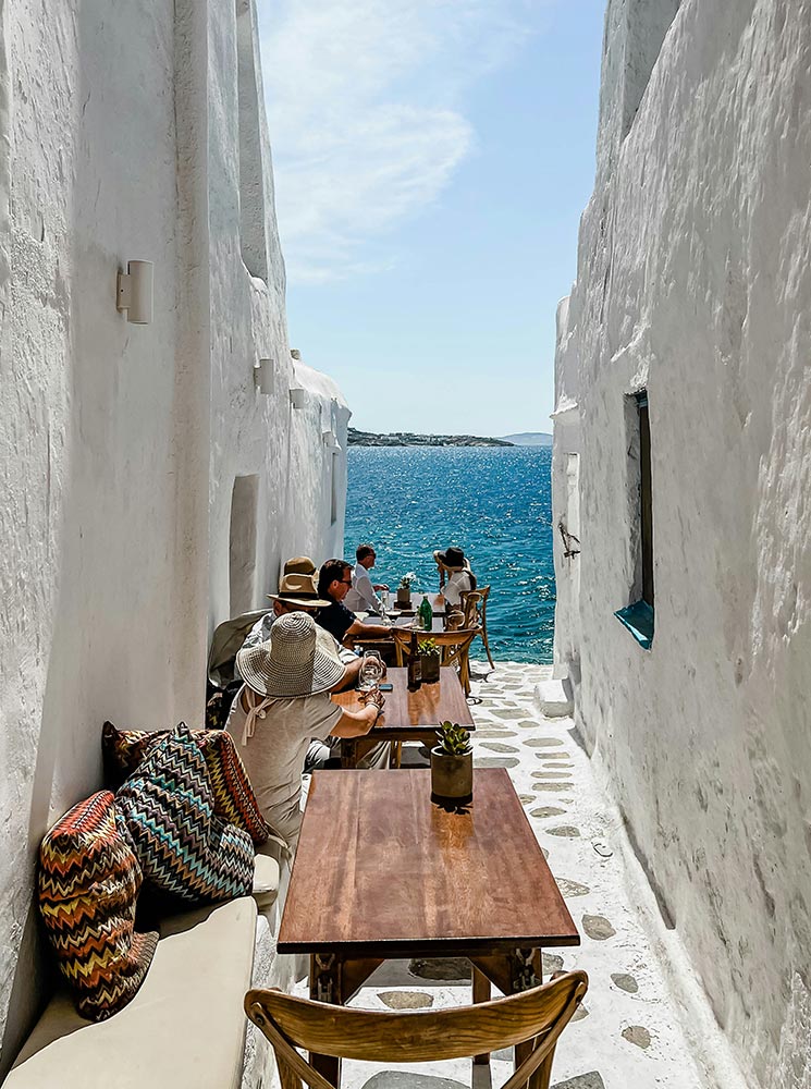 A narrow alleyway with whitewashed walls leads to a view of the Aegean Sea at Kastro’s restaurant in Mykonos. Wooden tables and chairs with colorful cushions line the path, where diners enjoy a sunny seaside meal.