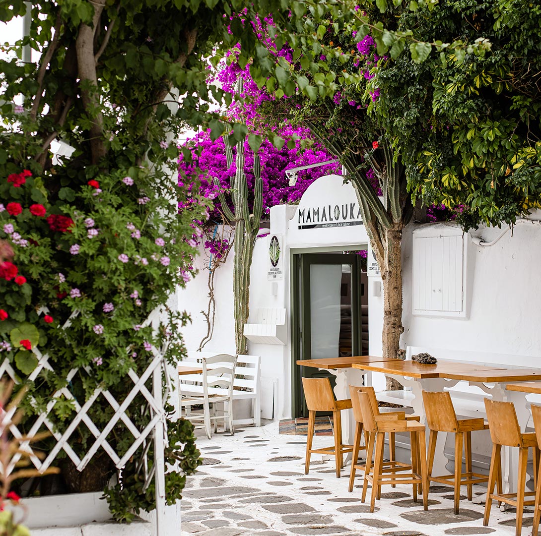 A charming outdoor seating area of Mamalouka restaurant in Mykonos, surrounded by vibrant purple bougainvillea, green foliage, and whitewashed walls. Wooden tables and chairs sit on a traditional cobblestone path, creating a picturesque and inviting atmosphere.