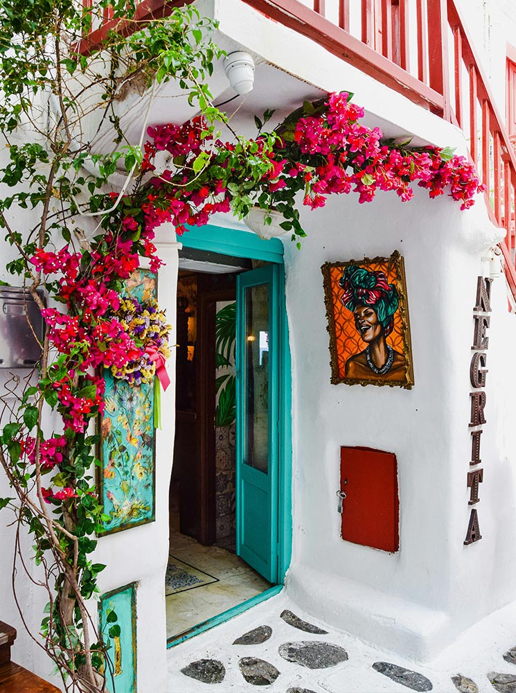 The entrance of Negrita Bar in Mykonos, featuring a vibrant turquoise door surrounded by pink bougainvillea flowers, colorful wall art, and the bar’s name displayed vertically on the whitewashed facade.