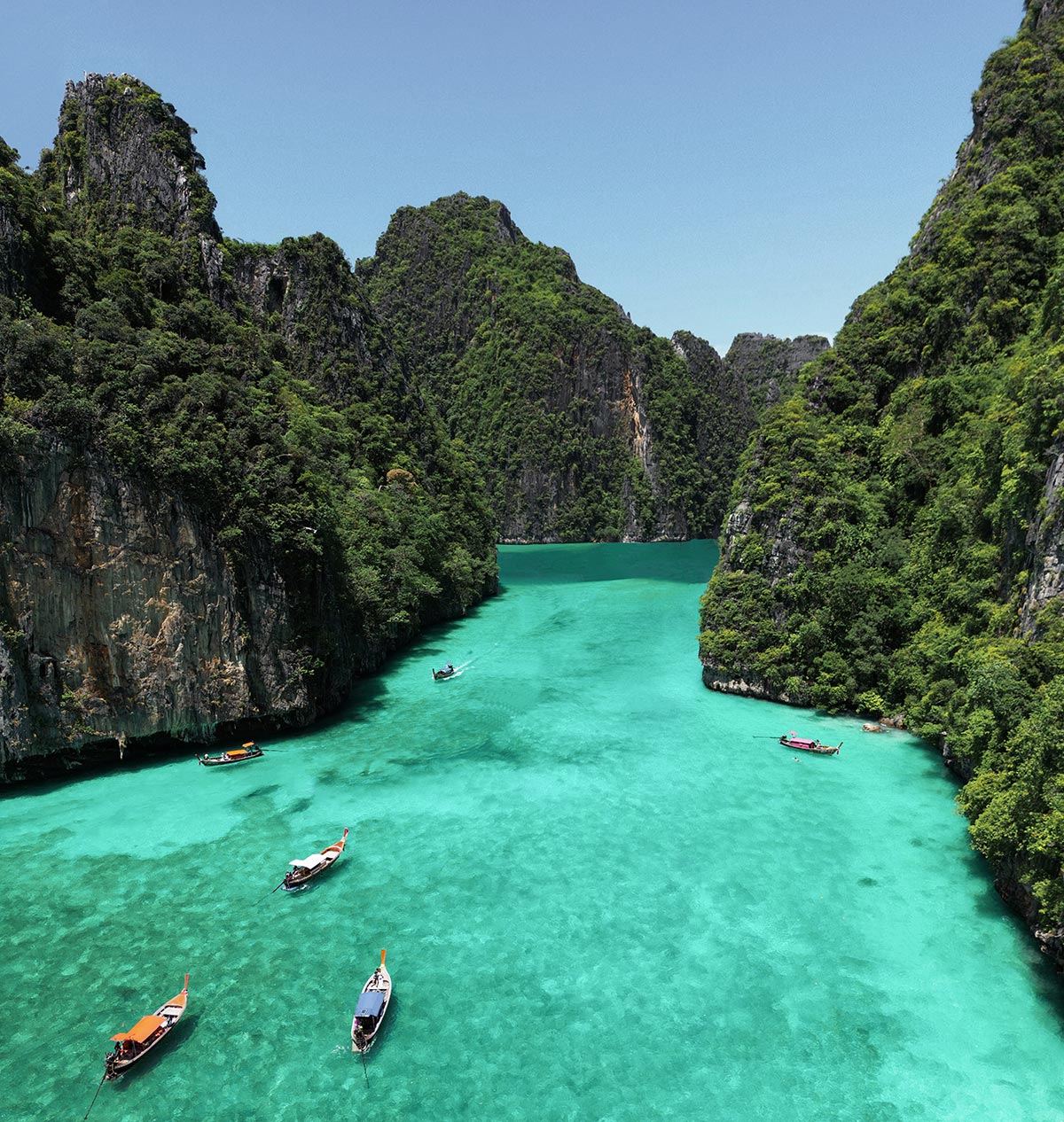 A breathtaking aerial view of turquoise waters surrounded by towering limestone cliffs covered in lush greenery, with traditional longtail boats scattered across the lagoon at the Phi Phi Islands.