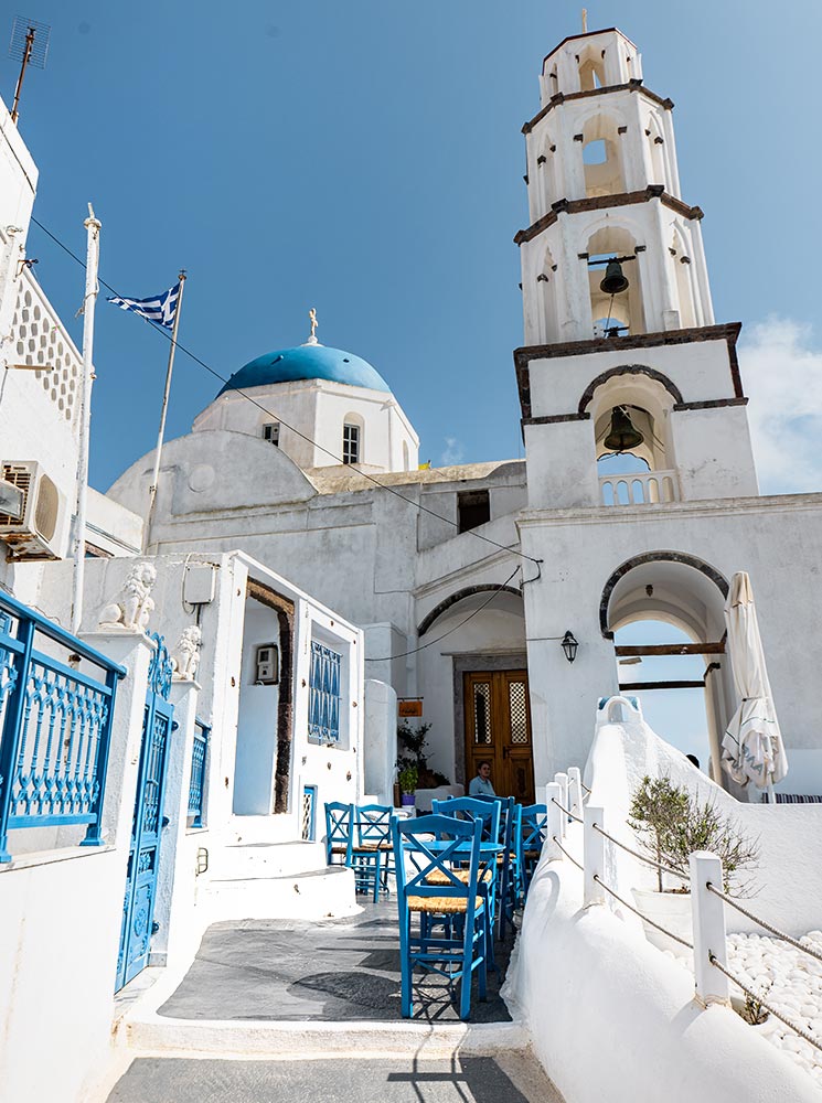 A traditional Greek church in Pyrgos, Santorini, featuring a white bell tower with dark accents, a blue-domed roof, and a courtyard with vibrant blue chairs and railings, set against a bright blue sky.