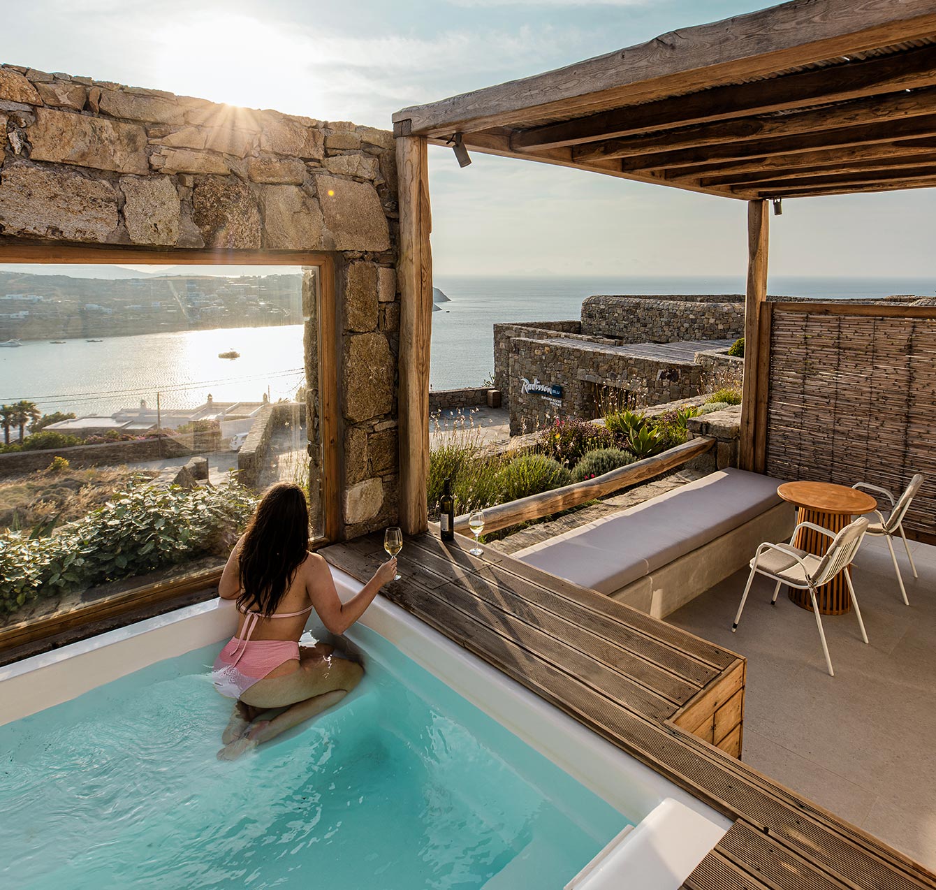 A relaxing view at Radisson Blu Euphoria Resort in Mykonos, featuring a woman in a pink swimsuit enjoying a private pool with a glass of wine, overlooking a scenic coastline and sparkling sea as the sun sets. The outdoor area includes wooden accents, comfortable seating, and natural stone walls, creating a tranquil ambiance.