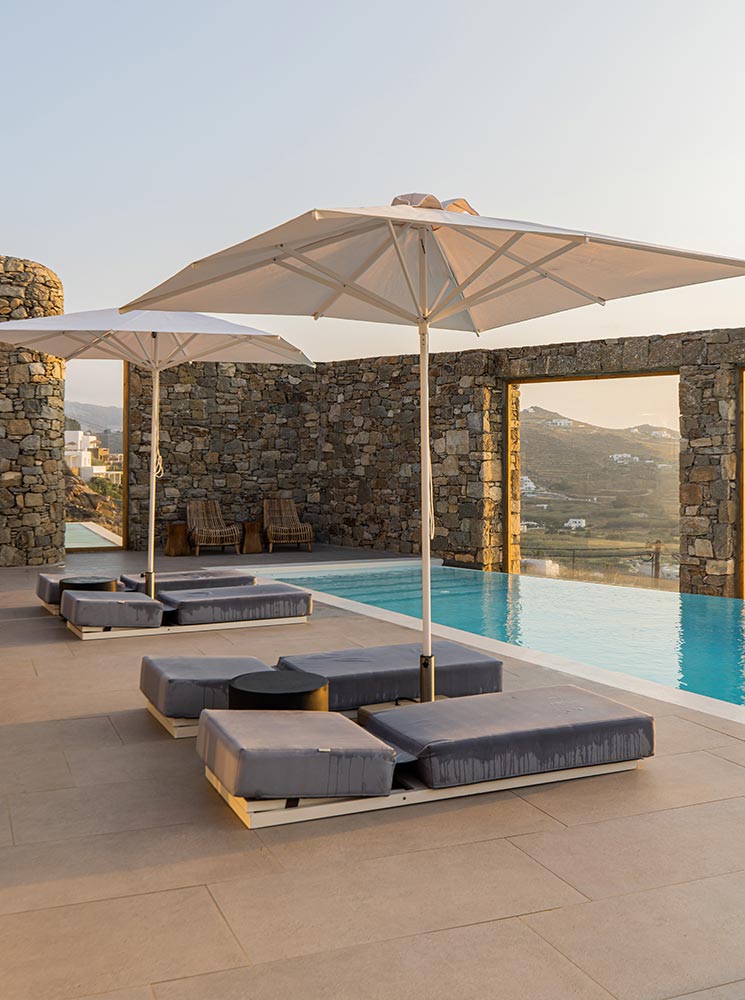 Poolside loungers with modern gray cushions and white umbrellas beside a serene infinity pool at the Radisson Blu Euphoria Resort in Mykonos. The stone wall frames the view of rolling hills and the golden sunset, creating a tranquil and luxurious atmosphere.