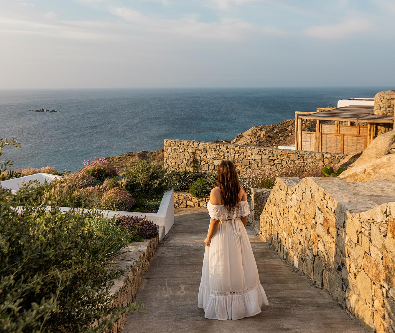 A woman in a flowing white dress walks down a stone-lined path, surrounded by lush greenery, towards the open sea at sunset. The Radisson Blu Euphoria Resort in Mykonos offers breathtaking ocean views, with rustic stone walls and serene natural beauty framing the scene.